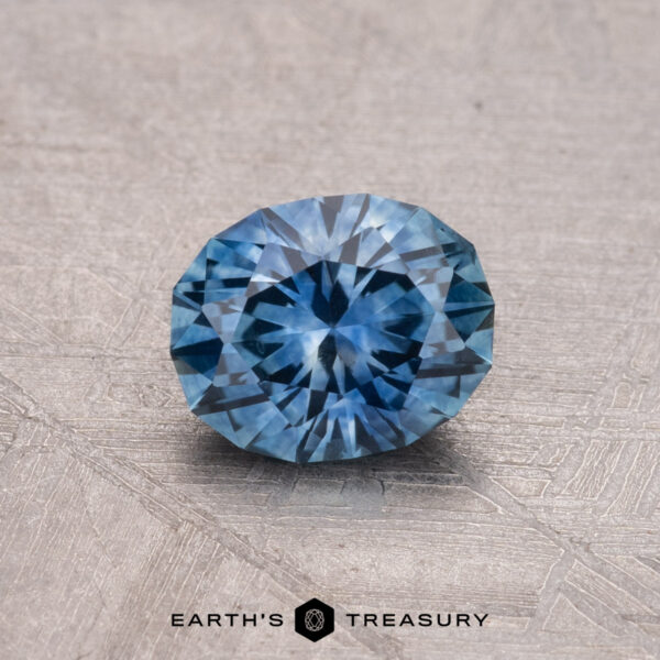 0.98-Carat Montana Sapphire (Heated) A round 0.98-carat heated Montana sapphire with intricate facets rests on a textured surface. Text reads: Earth’s Treasury.
