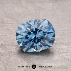 A 1.54-carat faceted round Montana sapphire gemstone, heated, on a textured surface with the "Earth's Treasury" logo.