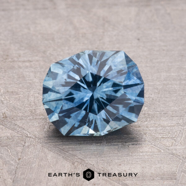 1.54-Carat Montana Sapphire (Heated) A 1.54-carat faceted round Montana sapphire gemstone, heated, on a textured surface with the "Earth's Treasury" logo.