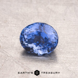 A 1.25-carat Ceylon sapphire gemstone, featuring a round blue shape, is placed on a textured surface, with the "Earth's Treasury" logo displayed at the bottom.