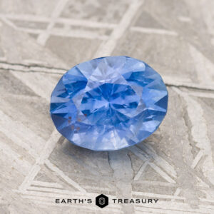 A 1.85-carat Cornflower Blue Ceylon Sapphire rests on a textured surface, with the "Earth's Treasury" logo at the bottom.