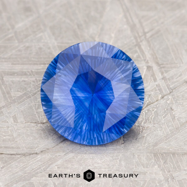 A 1.55-carat Ceylon sapphire with a faceted, round cut rests on a textured surface against a geometric-patterned background.