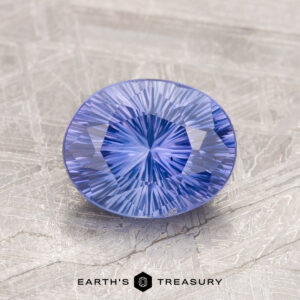 A 1.71-carat faceted blue Ceylon sapphire with intricate cuts sits on a textured surface, labeled as "EARTH'S TREASURY.