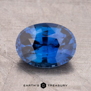 A 2.44-carat heated Ceylon sapphire, faceted in an oval shape, rests on a textured gray surface. Text reads "Earth's Treasury.