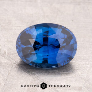 A 2.44-carat heated Ceylon sapphire, faceted in an oval shape, rests on a textured gray surface. Text reads "Earth's Treasury.