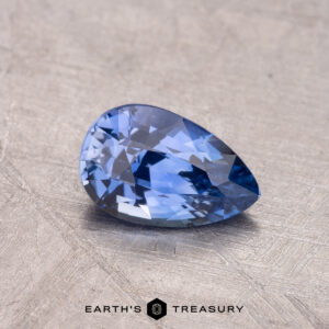 A 1.11-carat Ceylon sapphire, blue and faceted in a pear shape, rests on a textured surface with the "Earth's Treasury" logo beneath it.