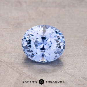 A 1.30-Carat Ceylon Sapphire, round and faceted in light blue, rests on a textured surface with "Earth's Treasury" labeled beneath.