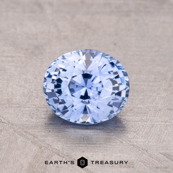 1.30-Carat Ceylon Sapphire A 1.30-Carat Ceylon Sapphire, round and faceted in light blue, rests on a textured surface with "Earth's Treasury" labeled beneath.