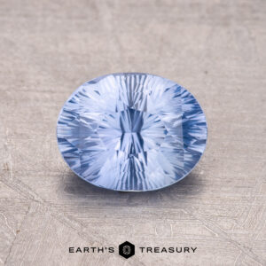 A 1.61-carat Ceylon Sapphire, faceted and round, features a light blue hue set against a textured background.