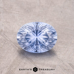 A faceted, oval-cut blue gemstone, the 1.47-Carat Ceylon Sapphire, rests on a textured surface with "Earth's Treasury" text visible at the bottom.