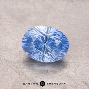 A blue oval-cut 1.34-carat Ceylon Sapphire gemstone is displayed on a textured surface with "Earth's Treasury" text at the bottom.