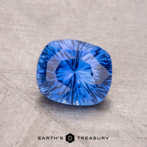 A vibrant blue 1.45-Carat Ceylon Sapphire with intricate facets lies on a textured surface, with "Earth's Treasury" visible below.