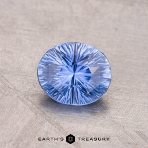 A 1.28-carat round Ceylon Sapphire, light blue in color with faceted detailing, displayed on a textured surface.