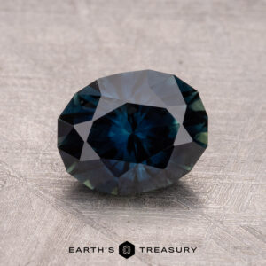 A 1.85-carat Australian sapphire (heated) in dark blue with facets lies on a textured light gray surface, labeled "Earth's Treasury.