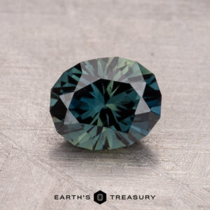 A faceted, blue-green gemstone with a textured surface, identified as a 1.53-carat Australian sapphire (heated), bears the inscription "Earth's Treasury.