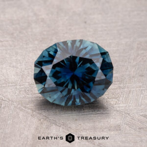 A round, faceted bluish gemstone glistens on a textured surface, etched with "Earth's Treasury." It is a 1.52-carat Australian Sapphire (Heated).