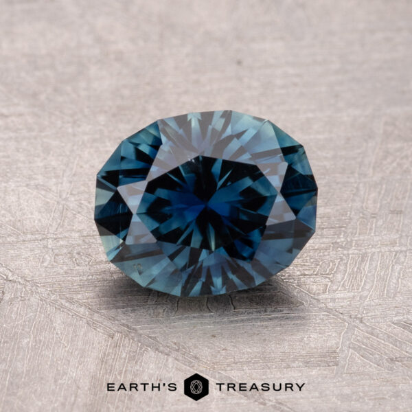 1.52-Carat Australian Sapphire (Heated) A round, faceted bluish gemstone glistens on a textured surface, etched with "Earth's Treasury." It is a 1.52-carat Australian Sapphire (Heated).