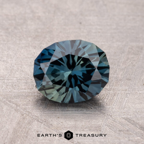1.41-Carat Australian Sapphire (Heated) A round, faceted gemstone from Earth's Treasury rests on a textured surface. It's a 1.41-carat, heated Australian sapphire with a captivating blue-green color.