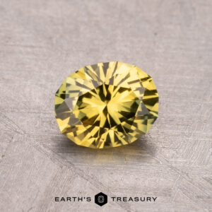 A 1.21-carat Australian Sapphire (Heated) gemstone, in a round cut with a yellow hue, rests on a textured gray surface with the text "Earth's Treasury" below.