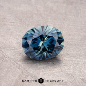 A 1.15-carat Australian Sapphire (Heated) with intricate facets rests on a textured surface, displaying the words "Earth's Treasury" at the bottom.