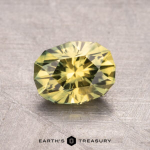 A 1.59-carat faceted yellow-green Australian sapphire (heated) on a textured gray surface with the "Earth's Treasury" logo at the bottom.