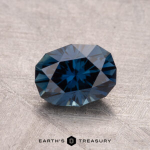 A faceted 1.55-carat heated Australian sapphire in dark blue with geometric cuts rests on a textured surface, marked by the "Earth's Treasury" logo below.