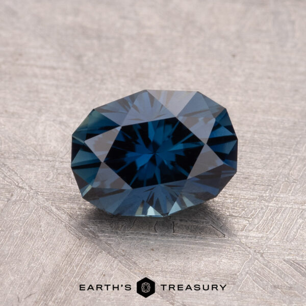 1.55-Carat Australian Sapphire (Heated) A faceted 1.55-carat heated Australian sapphire in dark blue with geometric cuts rests on a textured surface, marked by the "Earth's Treasury" logo below.