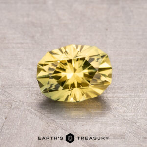 A 1.30-carat heated Australian sapphire, faceted and yellow, rests on a textured gray surface with the "Earth's Treasury" logo at the bottom.