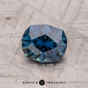 A faceted blue 1.32-carat Australian sapphire rests on a textured gray background. The text reads "Earth's Treasury" alongside a small geometric logo.