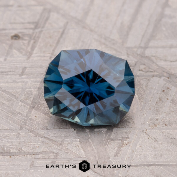 1.32-Carat Australian Sapphire (Heated) A faceted blue 1.32-carat Australian sapphire rests on a textured gray background. The text reads "Earth's Treasury" alongside a small geometric logo.