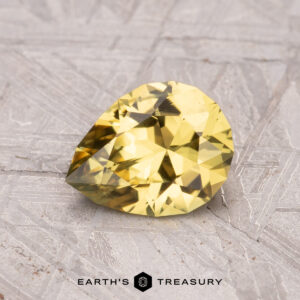 A 1.28-carat Australian sapphire (heated) in a pear shape rests on a textured gray surface, branded "Earth's Treasury" at the bottom.