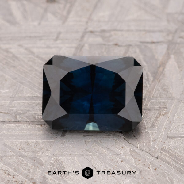 A 1.59-carat Australian Sapphire (heated), rectangular with faceted edges, rests on a textured gray surface.