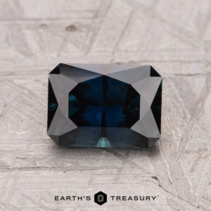 A rectangular, dark teal 1.55-carat Australian sapphire (heated) with facets is placed on a textured gray surface. Text below reads "Earth's Treasury.