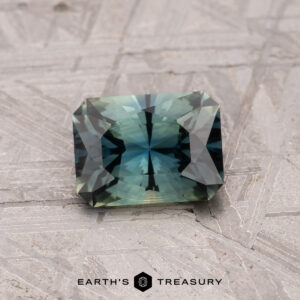 A 1.24-carat, rectangular bi-color Australian sapphire (heated), with blue and green hues, rests on a textured gray surface. The words "Earth's Treasury" appear at the bottom.