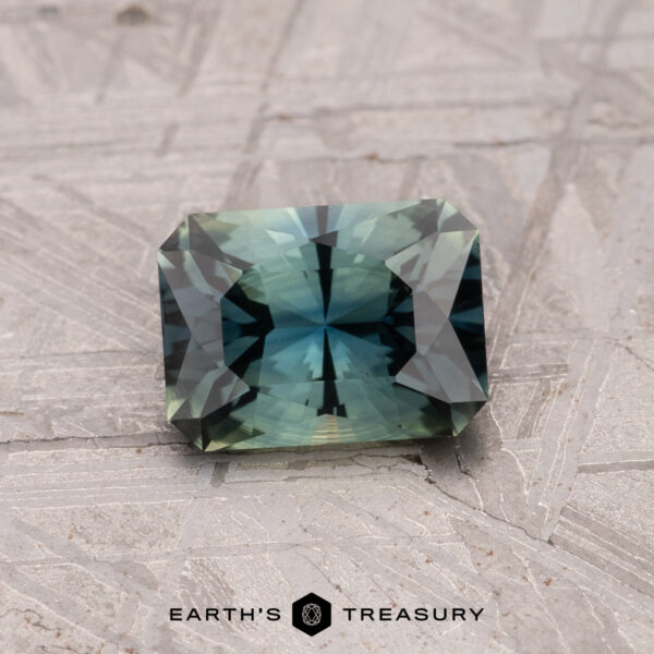 1.24-Carat Australian Sapphire (Heated) A 1.24-carat, rectangular bi-color Australian sapphire (heated), with blue and green hues, rests on a textured gray surface. The words "Earth's Treasury" appear at the bottom.