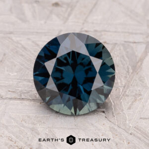 A round, faceted 1.99-carat Australian teal sapphire (heated) rests on a textured, light surface with "Earth's Treasury" at the bottom.