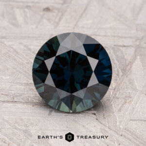 A round faceted dark gemstone, the 1.95-Carat Australian Sapphire (Heated), rests on a textured surface, with "Earth's Treasury" below it.