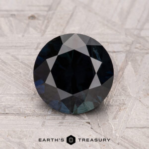 A 1.91-carat Australian sapphire, heated and dark blue with geometric facets, rests on a textured light gray surface. The "Earth's Treasury" logo is at the bottom.