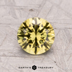 1.62-Carat Australian Sapphire (Heated), a round yellow gemstone with intricate facets on a textured gray surface, featuring the "Earth's Treasury" logo below.