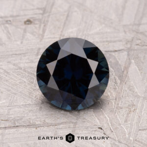 A round, faceted dark blue gemstone displayed on a textured surface with "Earth's Treasury" at the bottom is a 1.35-Carat Australian Sapphire (Heated).