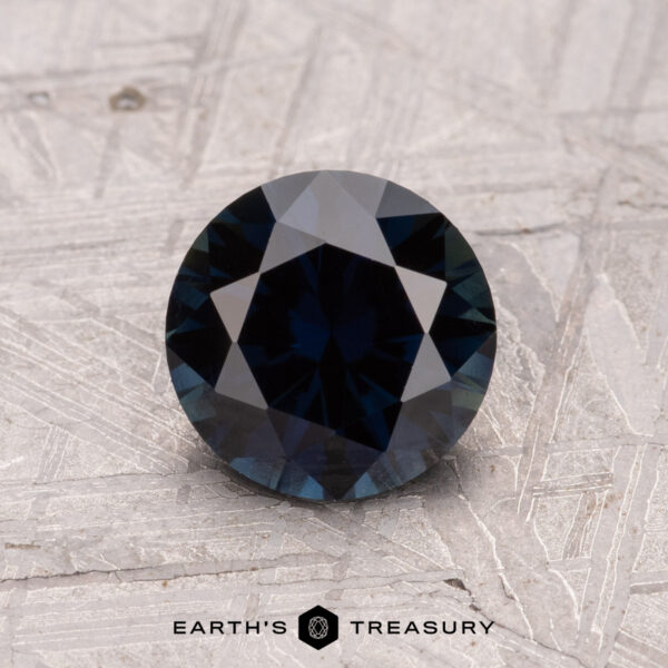 1.35-Carat Australian Sapphire (Heated) A round, faceted dark blue gemstone displayed on a textured surface with "Earth's Treasury" at the bottom is a 1.35-Carat Australian Sapphire (Heated).