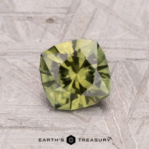 A square, green gemstone placed on a textured surface with the label "1.10-Carat Australian Sapphire (Heated)" underneath.