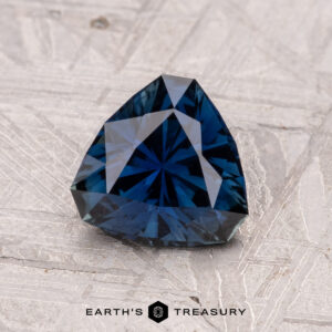 A faceted 1.16-carat Australian sapphire, with a dark blue color in a triangular shape, set against a textured gray background.