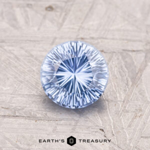 A 1.06-carat round, faceted blue Ceylon Sapphire with intricate patterns rests on a textured surface, featuring the "EARTH'S TREASURY" logo and text below.