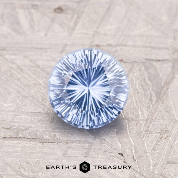 1.06-Carat Ceylon Sapphire A 1.06-carat round, faceted blue Ceylon Sapphire with intricate patterns rests on a textured surface, featuring the "EARTH'S TREASURY" logo and text below.