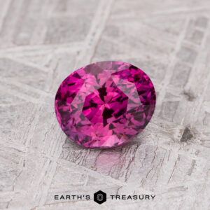 The 1.13-Carat Magenta Pink Ceylon Sapphire, an oval-cut gemstone, displayed on a textured gray surface with the "Earth's Treasury" logo below.