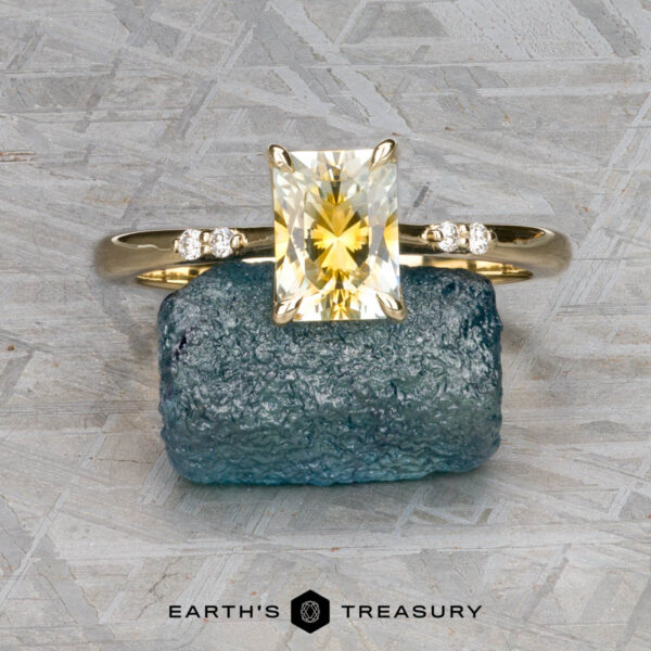 The "Kyria" ring in 14k yellow gold with 1.41-carat Montana sapphire The "Kyria" ring in 14k yellow gold with a 1.41-carat Montana sapphire is displayed on a rough blue-green stone, flanked by diamonds, against a patterned background with the "Earth's Treasury" logo below.