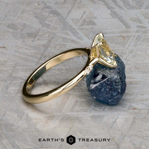 The "Kyria" ring in 14k yellow gold with 1.41-carat Montana sapphire The "Kyria" ring in 14k yellow gold with a 1.41-carat Montana sapphire is displayed against a rough blue stone on a textured gray surface, with the "Earth's Treasury" logo at the bottom.