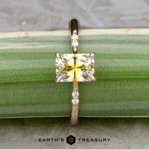 The "Kyria" ring is crafted in 14k yellow gold, featuring a 1.41-carat Montana sapphire and small round diamonds on each side, displayed against a green striped background.
