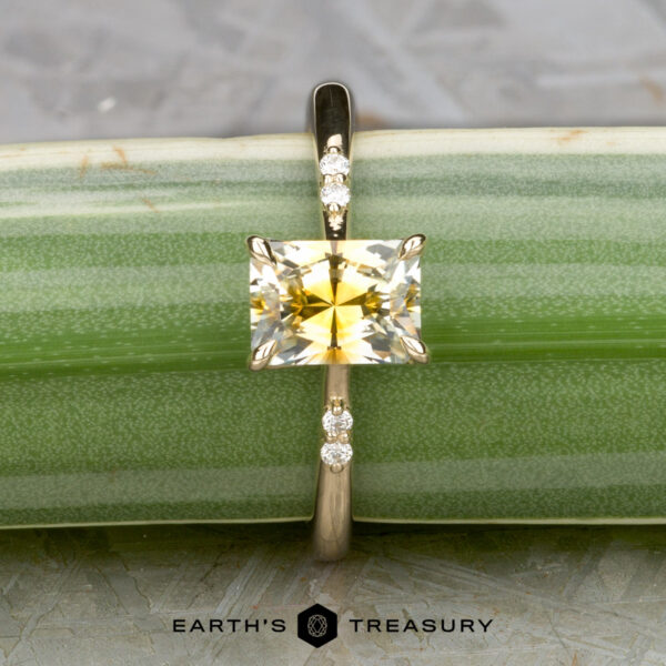 The "Kyria" ring in 14k yellow gold with 1.41-carat Montana sapphire The "Kyria" ring is crafted in 14k yellow gold, featuring a 1.41-carat Montana sapphire and small round diamonds on each side, displayed against a green striped background.
